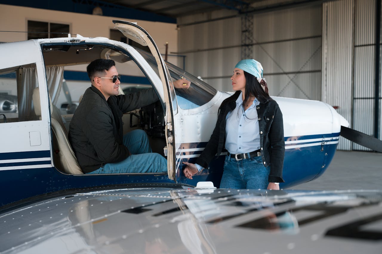 Man and woman interacting near a small plane in a hangar setting, discussing aviation.